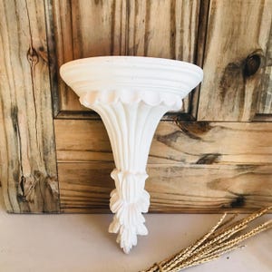 May include: A white, ornate wall shelf with a scalloped edge and decorative floral accents. The shelf has a flared, column-like support. The background is a wooden wall. The shelf is approximately 10 inches wide.