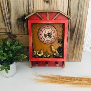 May include: A red, farmhouse-style wall clock with a chicken-themed design. The clock face is visible behind a wire mesh, and the frame has a small shelf with three hooks. The clock features a decorative roof and a painted finish.