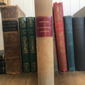 May include: A tan-colored book with a red spine and the title "STIEGLER LE MARECHAL OUDINOT" printed on the spine. The book is on a wooden shelf with other books.