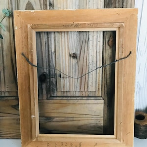 May include: A rectangular wooden picture frame shown from the back, featuring a light brown natural wood finish. A thin, dark metal hanging wire is attached to the frame sides. Handwritten text in black ink is visible on the top and bottom edges.
