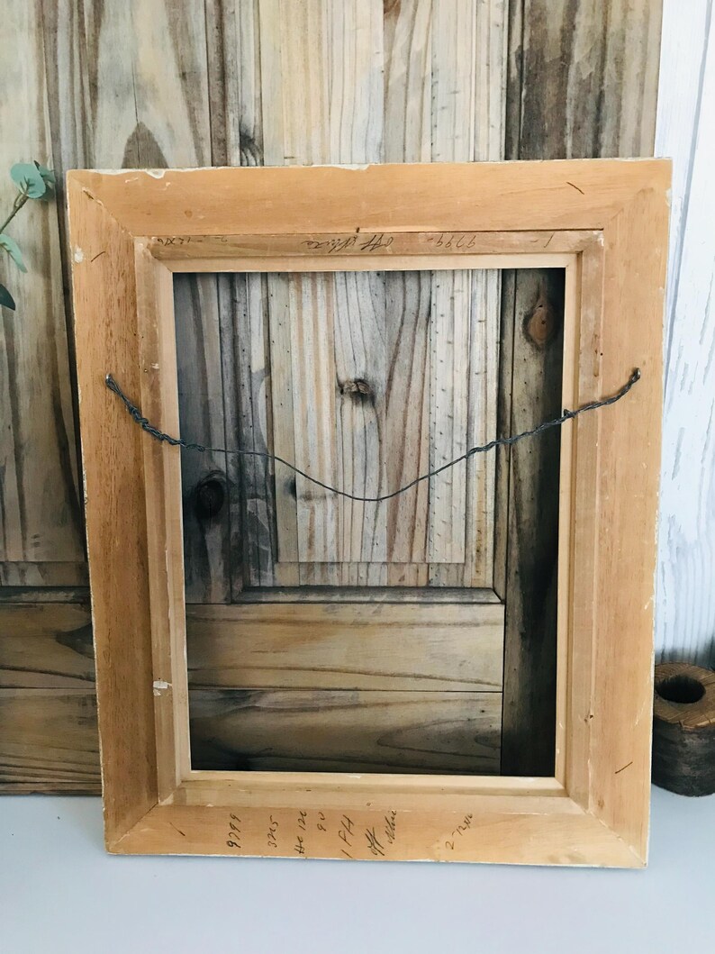May include: A rectangular wooden picture frame shown from the back, featuring a light brown natural wood finish. A thin, dark metal hanging wire is attached to the frame sides. Handwritten text in black ink is visible on the top and bottom edges.