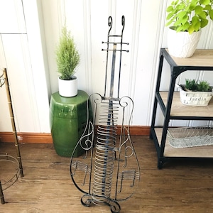 May include: A decorative metal magazine rack shaped like a cello. The rack is black with a spiral design for holding magazines. The cello-shaped rack has musical note accents and stands on a wooden floor.