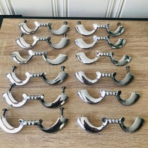 May include: A collection of polished silver-toned cabinet pulls arranged on a light wood surface. Each pull features a curved handle connected to a decorative centre piece and a screw post for installation. The pulls are displayed in neat rows.