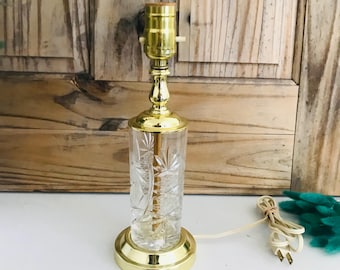 Op de afbeelding: Een gouden en heldere glazen tafellamp met een geslepen glasontwerp. De lamp heeft een gouden voet en een heldere glazen kap met een geslepen glasontwerp. De lamp staat op een wit oppervlak.