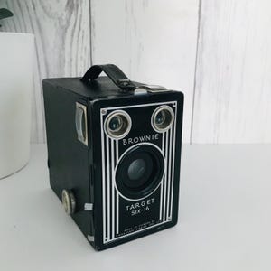 May include: A black vintage Brownie Target Six-16 camera with a silver trim and the words "Brownie" and "Target Six-16" printed on the front. The camera has a round lens and two small round viewfinders.