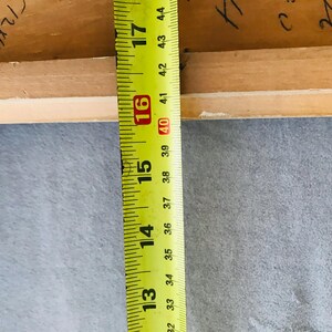 May include: A light brown wooden frame edge positioned next to a yellow measuring tape. The tape shows a measurement extending from 32 cm to over 44 cm. Handwritten black ink markings are visible on the inner surface of the wood.