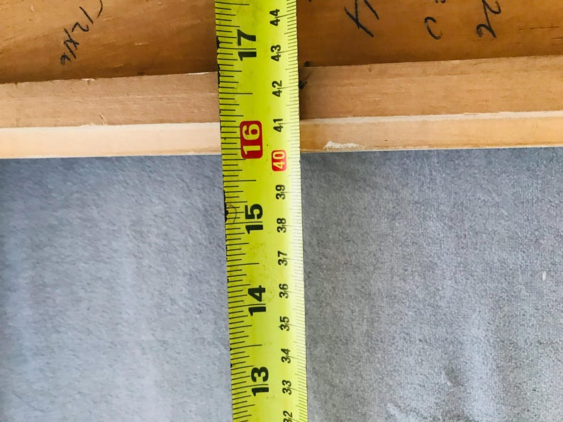 May include: A light brown wooden frame edge positioned next to a yellow measuring tape. The tape shows a measurement extending from 32 cm to over 44 cm. Handwritten black ink markings are visible on the inner surface of the wood.