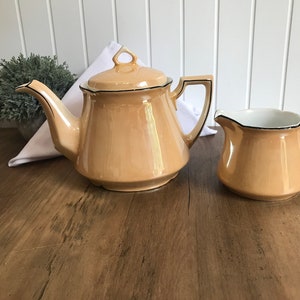 Op de afbeelding: Een vintage theeset met een crèmekleurige theepot en roomkan. Beide onderdelen hebben een zwarte rand rond de rand en een parelmoer afwerking.