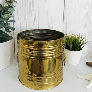 Könnte beinhalten: Ein goldfarbener Metalltopf mit geripptem Design und zwei dekorativen Griffen. Der Topf ist leer und steht auf einer weißen Oberfläche.