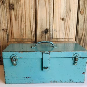 May include: A vintage, rectangular, turquoise metal toolbox with a handle and clasps. The toolbox has visible signs of wear and tear, suggesting age and use. The background features a wooden door with vertical planks.