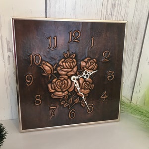 Peut inclure: Une horloge carrée en cuivre avec un cadre argenté et des chiffres romains pour les heures. Le cadran de l'horloge présente un motif en relief de trois roses.