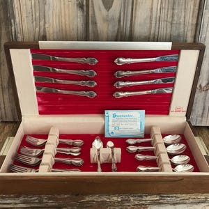 Puede incluir: Una caja de madera con un interior de terciopelo rojo que contiene un juego de cubiertos de plata. Los cubiertos incluyen cuchillos, tenedores y cucharas. La caja tiene una etiqueta que dice "Garantía".