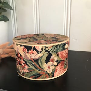 May include: A round, vintage floral box with a lid. The box is covered in a tapestry-like fabric featuring a pattern of pink, green, and cream-coloured flowers on a black background. The box is approximately 15 cm in diameter and 10 cm tall.
