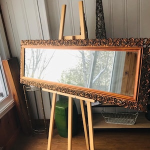 May include: A rectangular mirror with an ornate, bronze-colored frame. The mirror is propped on a wooden easel. The frame features detailed scrollwork. A green vase and a wire basket are visible in the background.