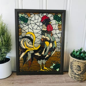 May include: A framed stained glass artwork featuring a colorful rooster. The rooster has a red comb and wattle, with yellow, black, brown, and gray feathers. Green grape clusters are in the background. The frame is dark brown.