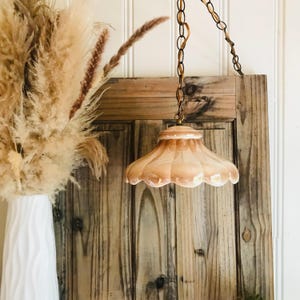 Op de afbeelding: Een hanglamp met een geschulpte, perzikkleurige glazen kap met een glanzende afwerking. De lamp hangt aan een donkere metalen ketting. Daarnaast staat een hoge, witte vaas met een golvende textuur en een groot boeket gedroogd pampasgras.
