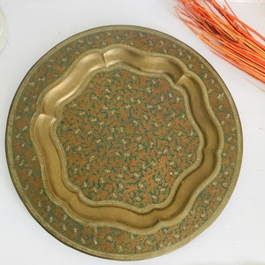 Op de afbeelding: Een ronde serveerschaal van messing met een decoratief ontwerp van groene en oranje bloemmotieven. De schaal heeft een verhoogde rand en een gegolfde rand.