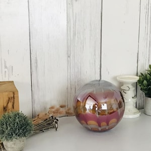 Puede incluir: Una esfera de vidrio decorativa con un acabado metálico. La esfera tiene un patrón de círculos superpuestos en tonos de rosa, naranja y dorado. La esfera está sentada sobre una superficie blanca.