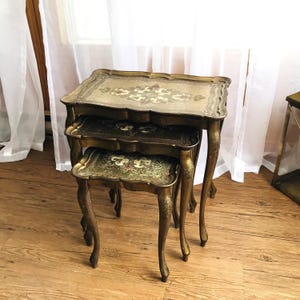 Puede incluir: Un juego de tres mesas nido antiguas con patas curvas ornamentadas y tableros decorativos. Las mesas tienen un acabado dorado con detalles florales y de volutas en tonos rojos, verdes y crema. Las mesas están dispuestas en una configuración anidada.