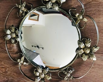 May include: A round, gold-toned mirror with a decorative floral frame. The mirror has a convex surface and is surrounded by stylised flower designs.