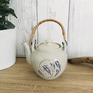 May include: A cream ceramic teapot with a bamboo handle and lid. The teapot has a design of purple leaves and brown stems. The teapot is on a wooden surface.