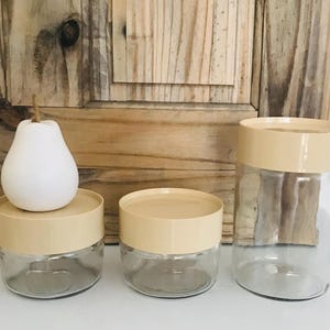 Peut inclure: Trois bocaux de rangement en verre avec des couvercles beiges. Les bocaux sont transparents et de forme ronde. Les couvercles sont lisses et ont un bord légèrement surélevé.