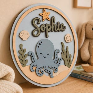 May include: A round wooden wall sign with a light blue background features an octopus, starfish, and the name "Sophie" in script. The sign has a light brown border and is decorated with sea-themed elements.