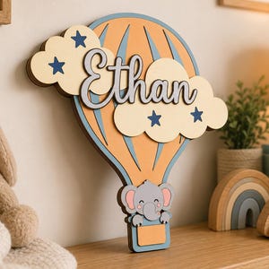 May include: A wooden hot air balloon-shaped wall decoration with the name "Ethan" in cursive. The balloon is orange and blue, with a cloud and star design. An elephant is in the basket. Decorative items are on a wooden surface.