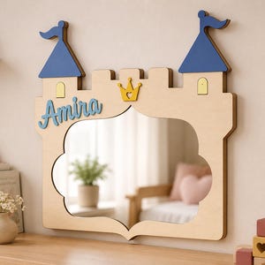 Puede incluir: Espejo de madera con forma de castillo con torres azules y una corona dorada. El nombre "Amira" está escrito en azul en el espejo. El espejo está sobre una cómoda de madera con juguetes y decoración.