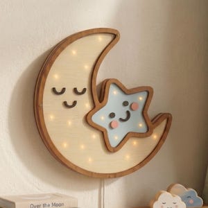 May include: Wooden crescent moon and star-shaped night light. The moon has a smiling face and is made of light-colored wood. The star is light blue with a smiling face and pink cheeks. The night light has small lights inside.