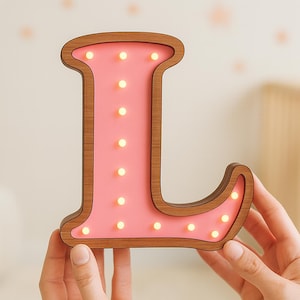 Op de afbeelding: Een houten letter "L" met een roze binnenkant en kleine, warm getinte lampjes. De letter heeft een bruin houten frame en wordt door een persoon vastgehouden. De achtergrond is een zachte, neutrale kleur, wat duidt op een kinderkamer.