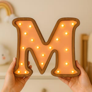 Puede incluir: Una letra "M" de madera con un interior color melocotón y pequeñas luces blancas cálidas. La letra se sostiene frente a un fondo neutro. La parte superior de la imagen dice "SVG DXF DWG AI PNG" y "INSTANT DOWNLOAD".