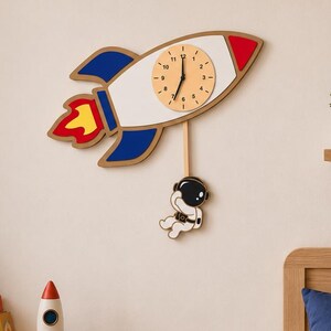 Op de afbeelding: Een houten wandklok in de vorm van een raket met blauwe, rode en witte accenten, en een hangende astronautfiguur. De wijzerplaat is beige met zwarte cijfers en wijzers. De kamer heeft ook een teddybeer, een stervormig kussen en een bed.