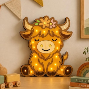 May include: A wooden light-up Highland cow with a flower crown, alongside a teddy bear, wooden train, and a rainbow stack of books. The books read "Little Explorer", "Dream Big", and "You Are Loved". A banner and framed landscape are also visible.
