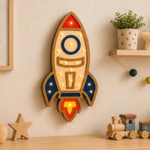 Peut inclure: Décoration murale en forme de fusée en bois, avec des accents rouges, bleus et jaunes. La fusée est éclairée et entourée d'autres décorations de chambre d'enfant, dont un ours en peluche, des étoiles et un train en bois. Le texte "INSTANT DOWNLOAD" est visible.