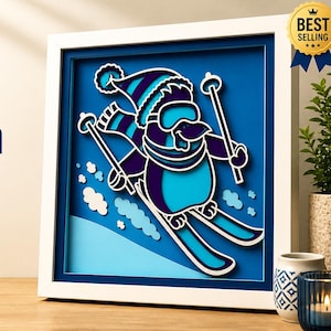 Peut inclure: Boîte à ombre multicouche avec un motif de pingouin skieur, découpé au laser, dans des tons de bleu et de violet. Le texte indique « Skiing Penguin » et « Mandala Decor ». Cadre blanc.