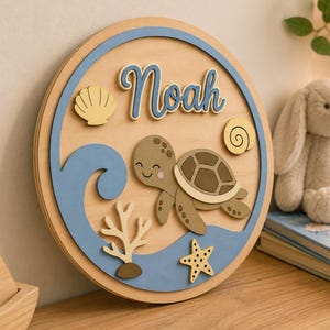 Puede incluir: Decoración de pared redonda de madera con un diseño de tortuga marina, ola y estrella de mar. El nombre "Noah" está escrito en azul. La obra de arte utiliza tonos marrones, azules y beige, con una concha y una espiral.