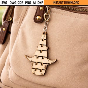 May include: A wooden keychain with a unique design, hanging from a beige bag. The keychain is made of light-colored wood and features a layered, abstract shape with horn-like extensions. The bag has brown leather accents and a zipper.