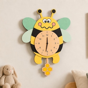 May include: A whimsical yellow and black bee-shaped wall clock with a light-colored clock face. The clock features a honeycomb pendulum and green wings. The clock is mounted on a light-colored wall, with a shelf, books, and a stuffed bunny in the background.