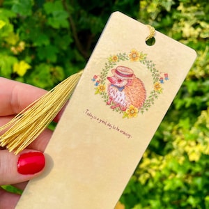 Könnte beinhalten: Beige Lesezeichen mit einem Cartoon-Igel mit Hut und Fliege, umgeben von einem Blumenkranz. Das Lesezeichen hat eine goldene Quaste und den Text "Today is a great day to be amazing."