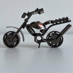May include: A small, metal motorcycle sculpture made from nuts, bolts, and other hardware. The motorcycle is painted a dark bronze color and has a detailed design.
