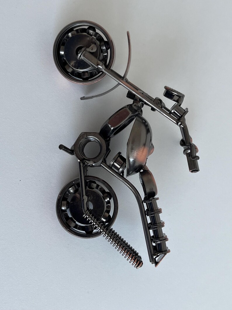 May include: A small, metal motorcycle sculpture made from nuts, bolts, and other hardware. The motorcycle is in a wheelie position with the front wheel in the air. The sculpture is painted in a dark metallic color.