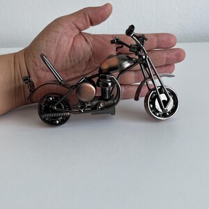 May include: A small, metal motorcycle figurine with a copper-colored gas tank and black metal accents. The motorcycle is on a white surface.