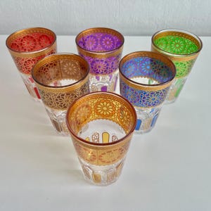 May include: Six small, clear glass cups with gold trim and intricate patterns. The cups are filled with different coloured liquids, including red, purple, green, and blue.
