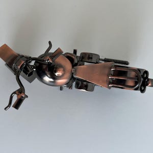 May include: A small, metal motorcycle sculpture with a copper finish. The motorcycle is made of various metal pieces, including a fork, a wrench, and a piece of wire mesh. The motorcycle is in a tilted position, with the front wheel facing the viewer.