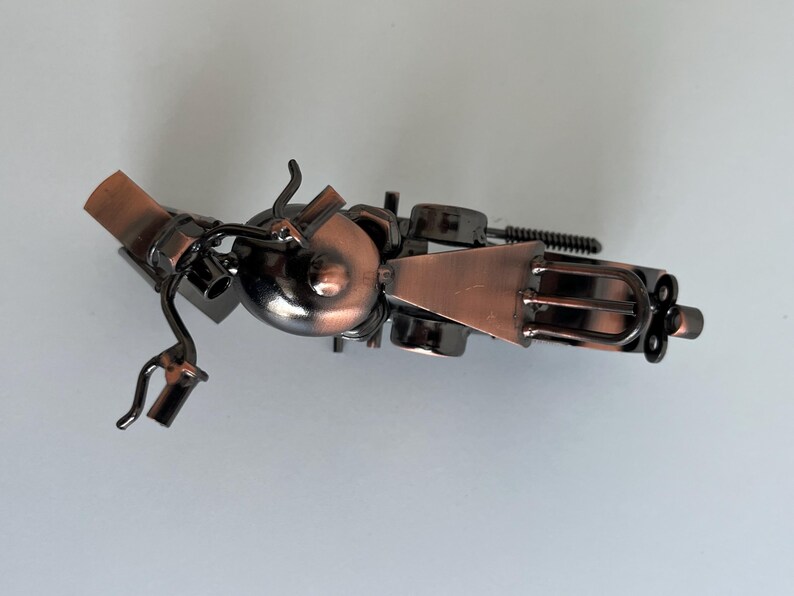 May include: A small, metal motorcycle sculpture with a copper finish. The motorcycle is made of various metal pieces, including a fork, a wrench, and a piece of wire mesh. The motorcycle is in a tilted position, with the front wheel facing the viewer.