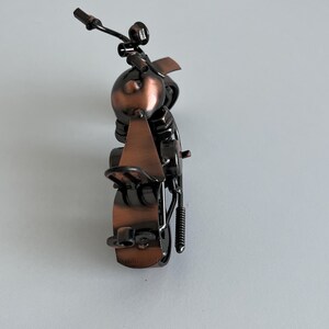 May include: A small, metal motorcycle sculpture with a copper finish. The motorcycle is made of various metal pieces, including nuts, bolts, and wire. The motorcycle is on a white surface.