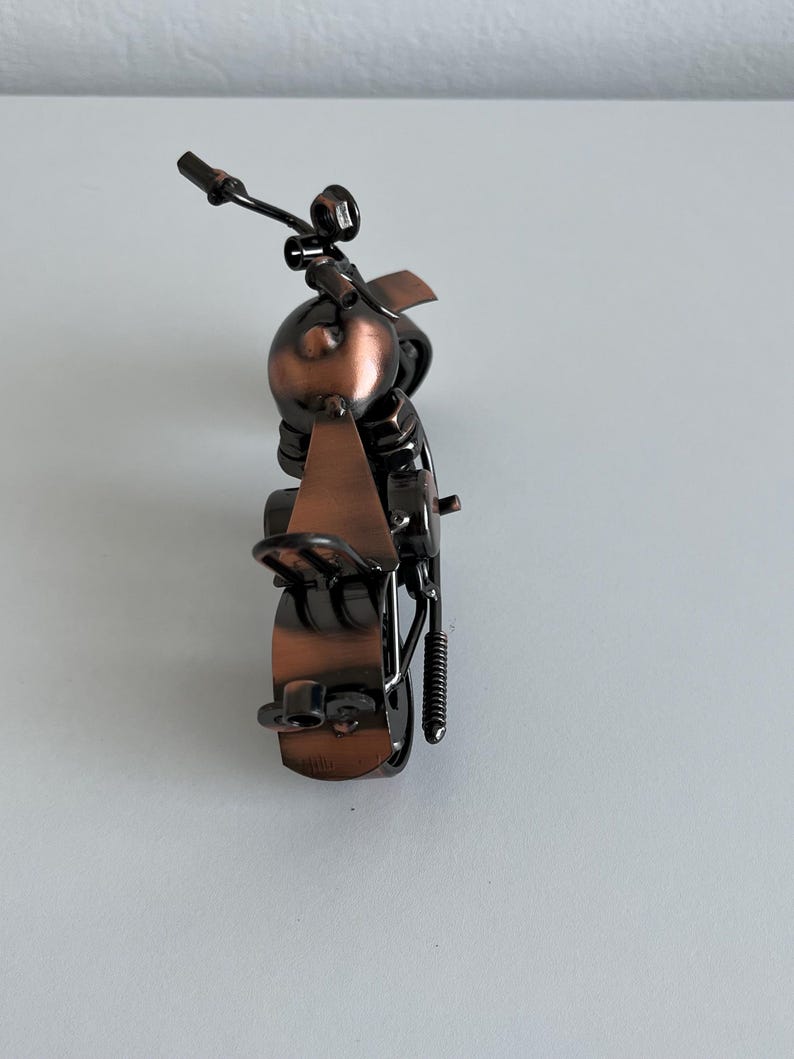 May include: A small, metal motorcycle sculpture with a copper finish. The motorcycle is made of various metal pieces, including nuts, bolts, and wire. The motorcycle is on a white surface.