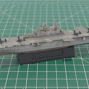 May include: A grey model aircraft carrier with a flat deck and miniature aircraft. The ship, displaying the number "6", is mounted on a black base. The model is set against a green grid background, with measurements in centimetres.