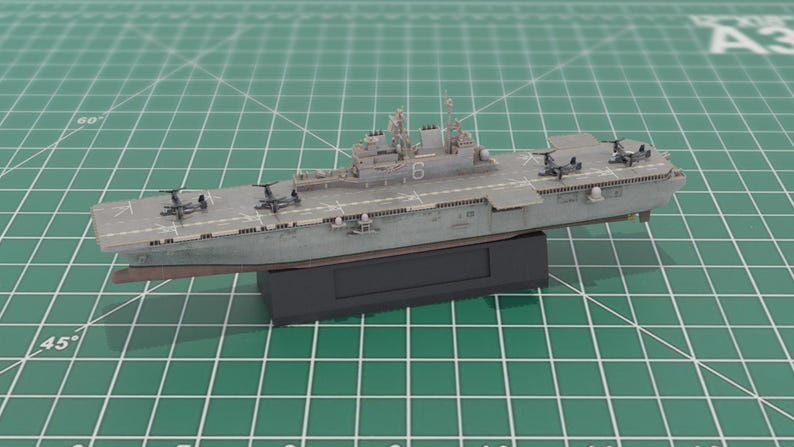 May include: A grey model aircraft carrier with a flat deck and miniature aircraft. The ship, displaying the number "6", is mounted on a black base. The model is set against a green grid background, with measurements in centimetres.